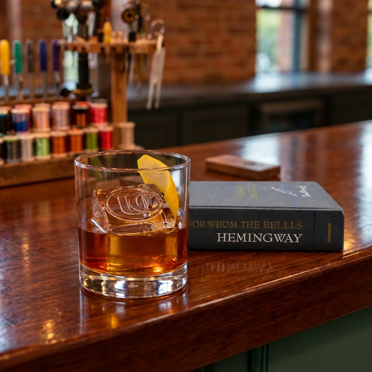 An Upland Club monogrammed ice cube in a whiskey glass beside a Hemingway novel on the mahogany bar, with fly-tying thread spools behind